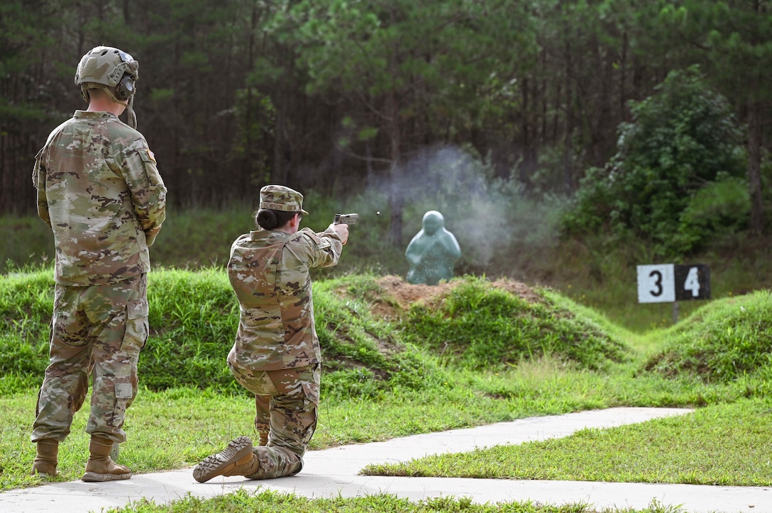 A guardsman takes a knee and fires a service weapon at a green target behind mounds of grass with a forest behind, while a guardsman stands to the left and watches.