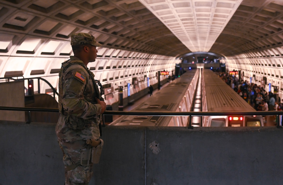 A guardsman stands on a balcony overlooking two trains in a station, as crowds of people walk below to the right.