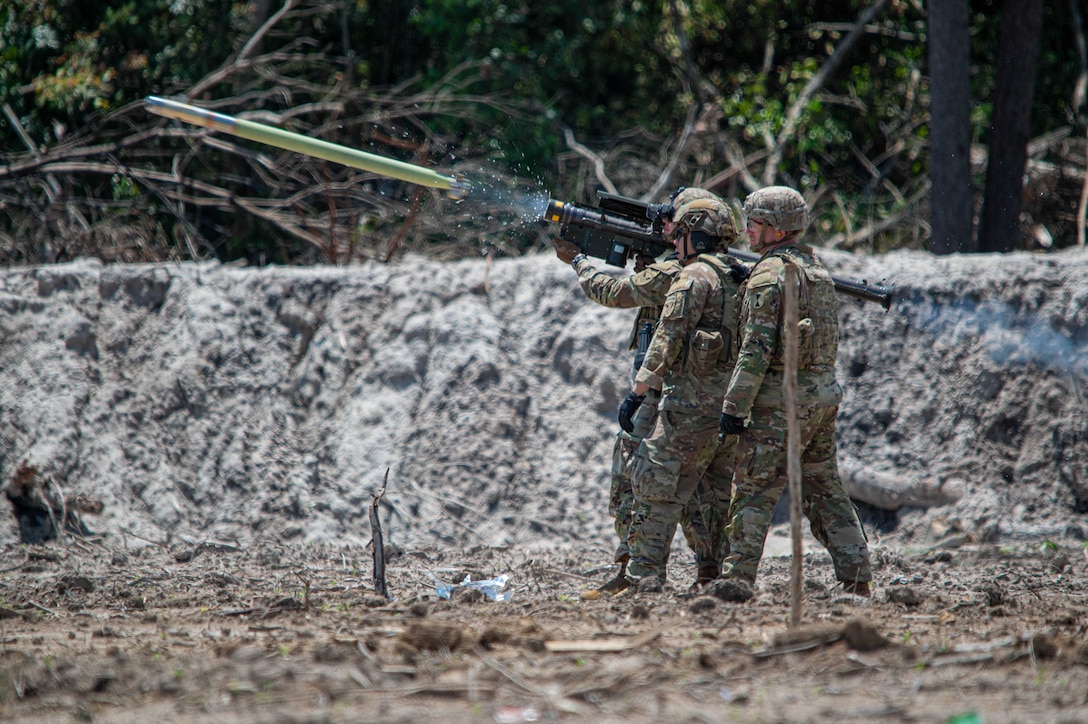 Three soldiers in helmets hold up a portable air defense system and fire a missile in a sandy area with brush behind.