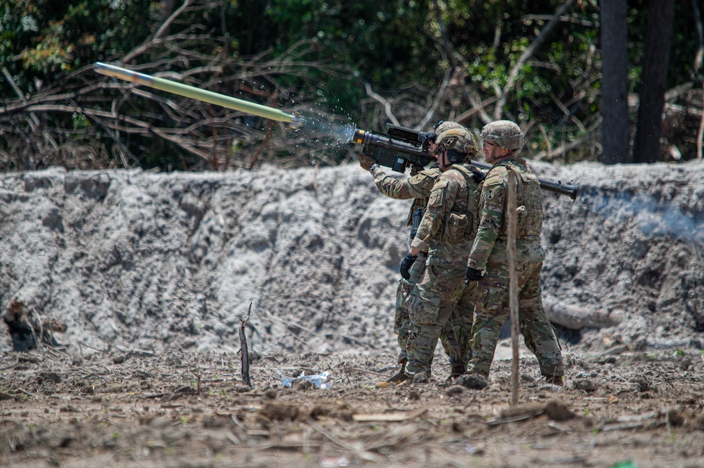 Three soldiers in helmets hold up a portable air defense system and fire a missile in a sandy area with brush behind.