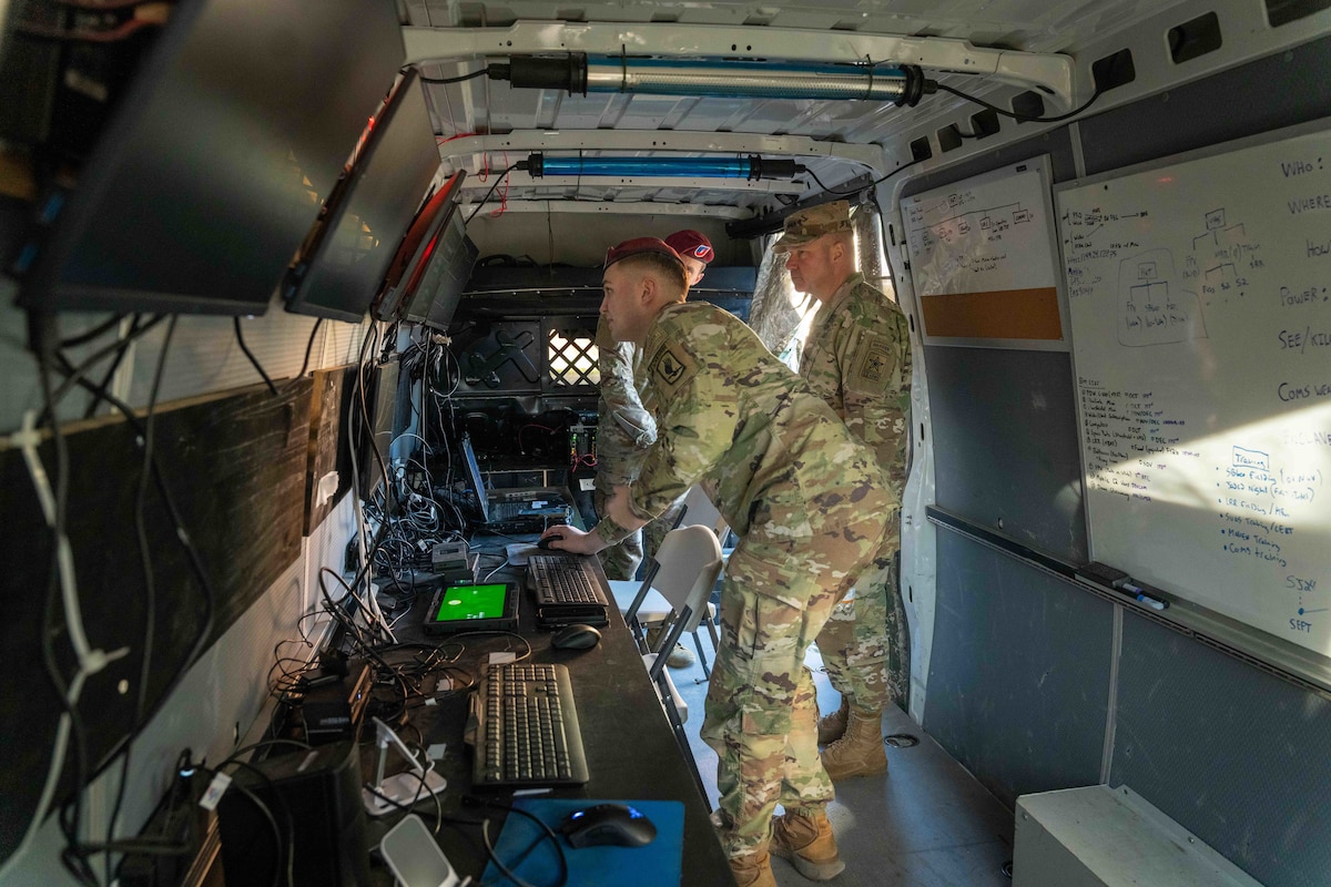 A man in a camouflage military uniform looks at a computer screen while moving a mouse. Another soldier in similar attire looks at the screen. Both men and another soldier in the background are standing in a long vehicle with a line of computers and monitors on the left side.
