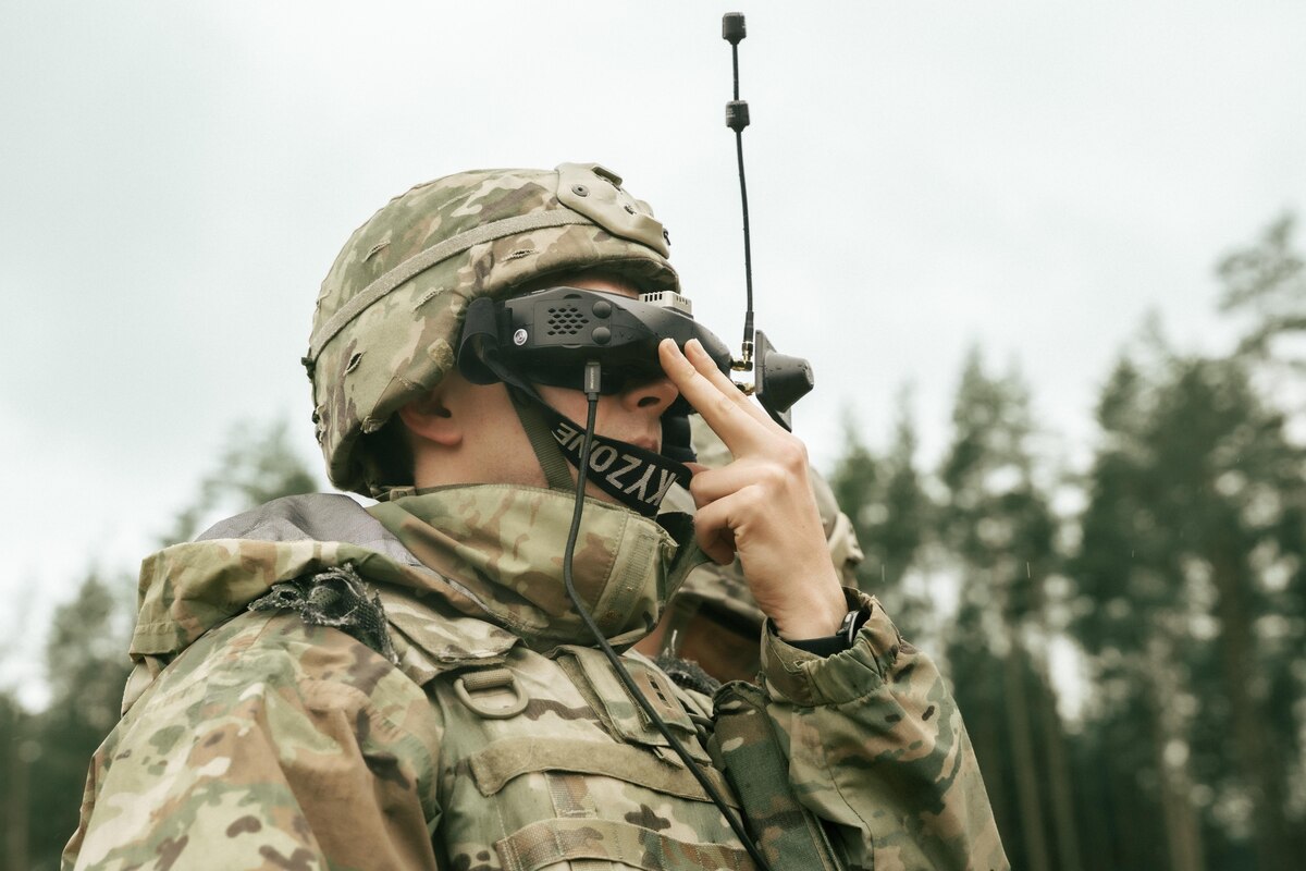 A soldier in a camouflage military uniform and helmet adjusts tactical goggles on his face.