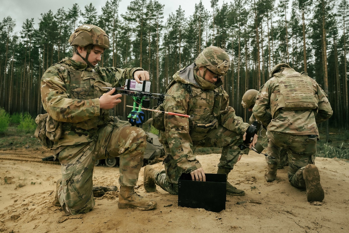 A group of soldiers in camouflage military uniforms kneel in the sand. A soldier on the left is holding a drone, while the soldier to the right is holding a remote. There are trees in the background.