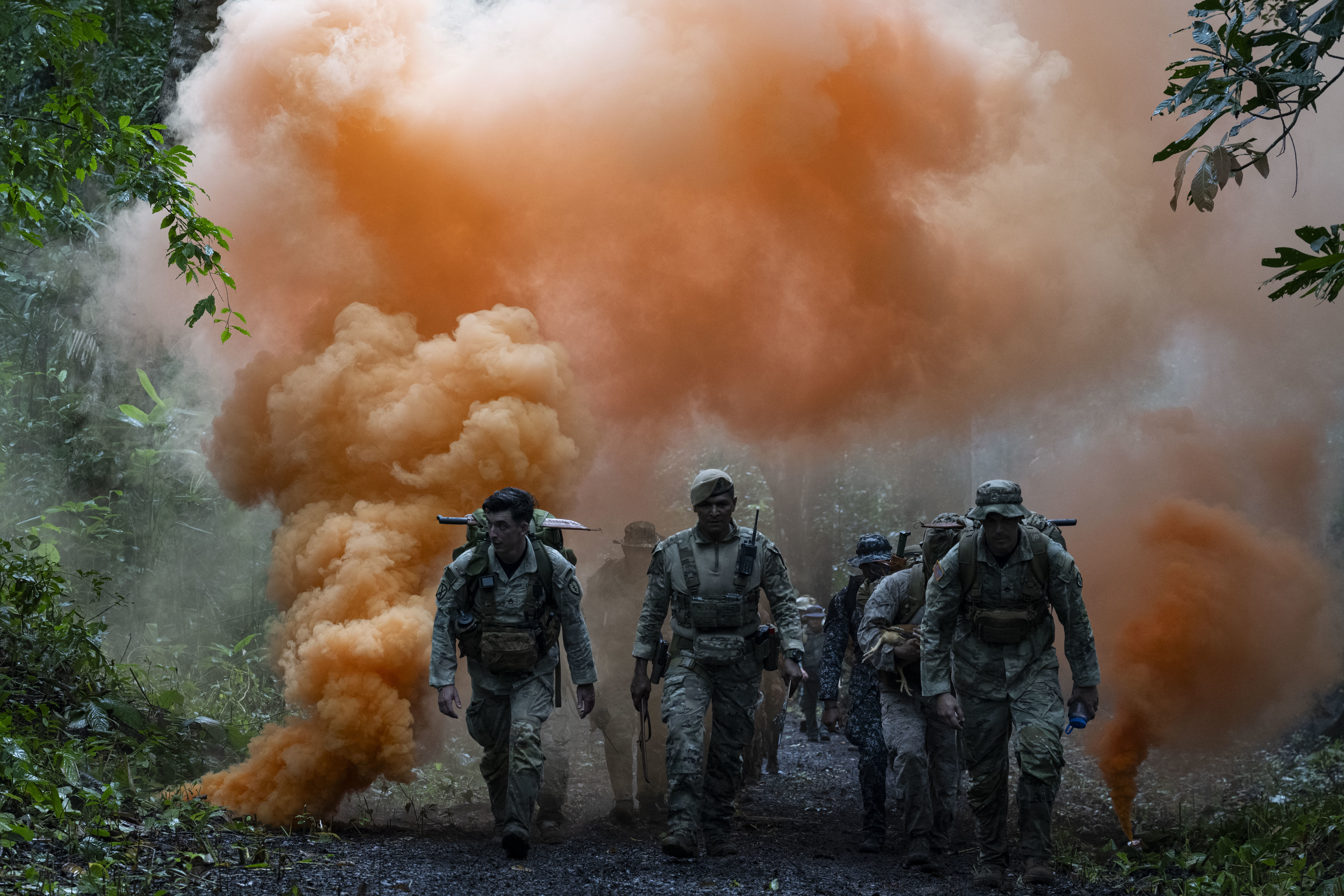Men in camouflage uniforms walk in the jungle with orange smoke rising behind them in the background. Men in camouflage uniforms walk in the jungle with orange smoke rising behind them in the background.