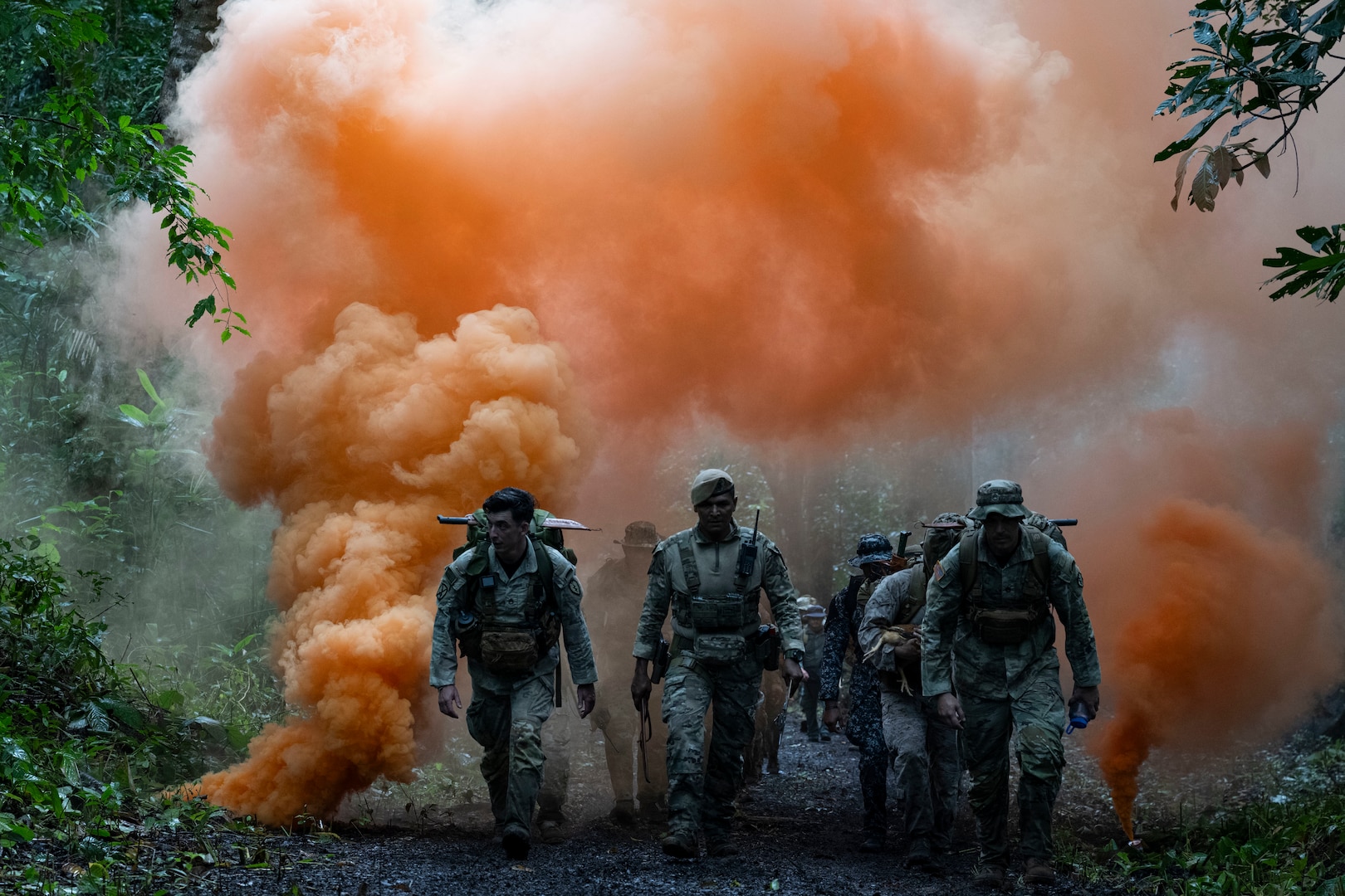Men in camouflage uniforms walk in the jungle with orange smoke rising behind them in the background.