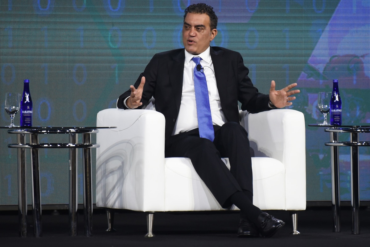 A person in a suit and blue tie sits in a seat and speaks, gesturing with both hands.