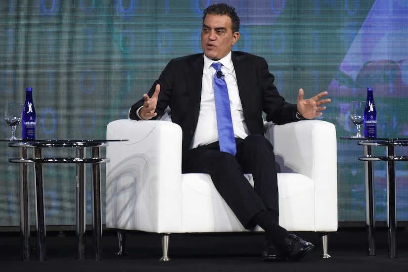 A person in a suit and blue tie sits in a seat and speaks, gesturing with both hands.
