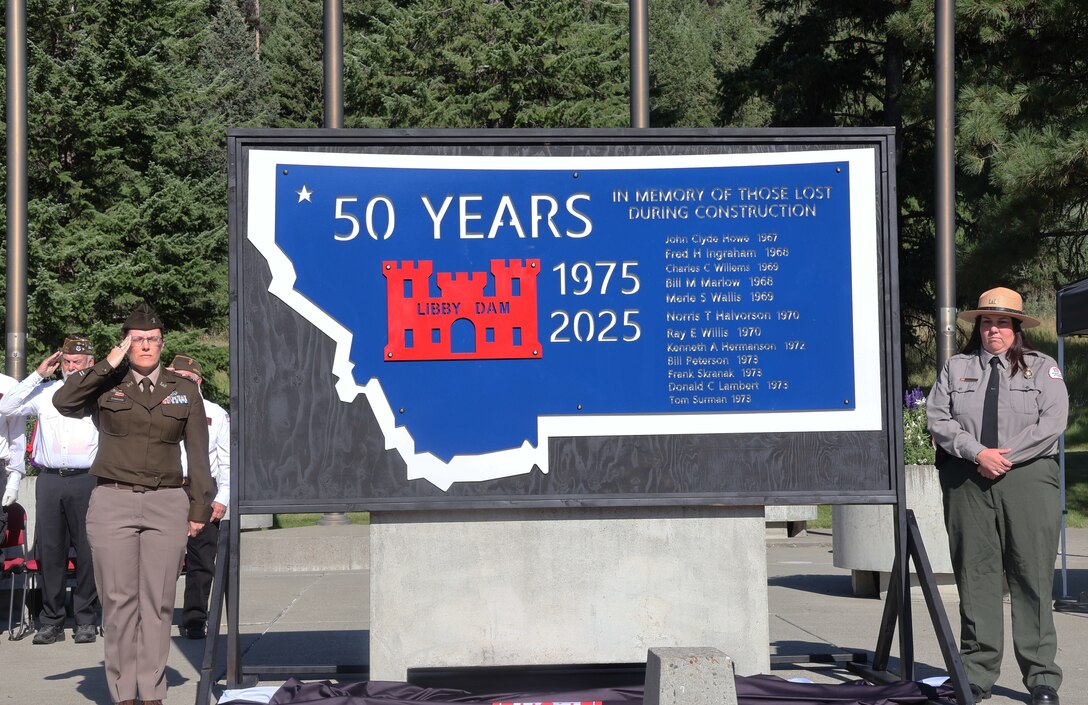U.S. Army Corps of Engineers, Seattle District Commander Col. Kathryn Sanborn and Libby Dam Natural Resource Manager Tana Wilson stand at attention while the VFW and American Legion perform Taps and the memorial unveiling, during Libby Dam's 50th commemoration, Aug. 23, 2025. The memorial lists the names of the 12 men who lost their lives during the construction of the dam between 1966 and 1972.