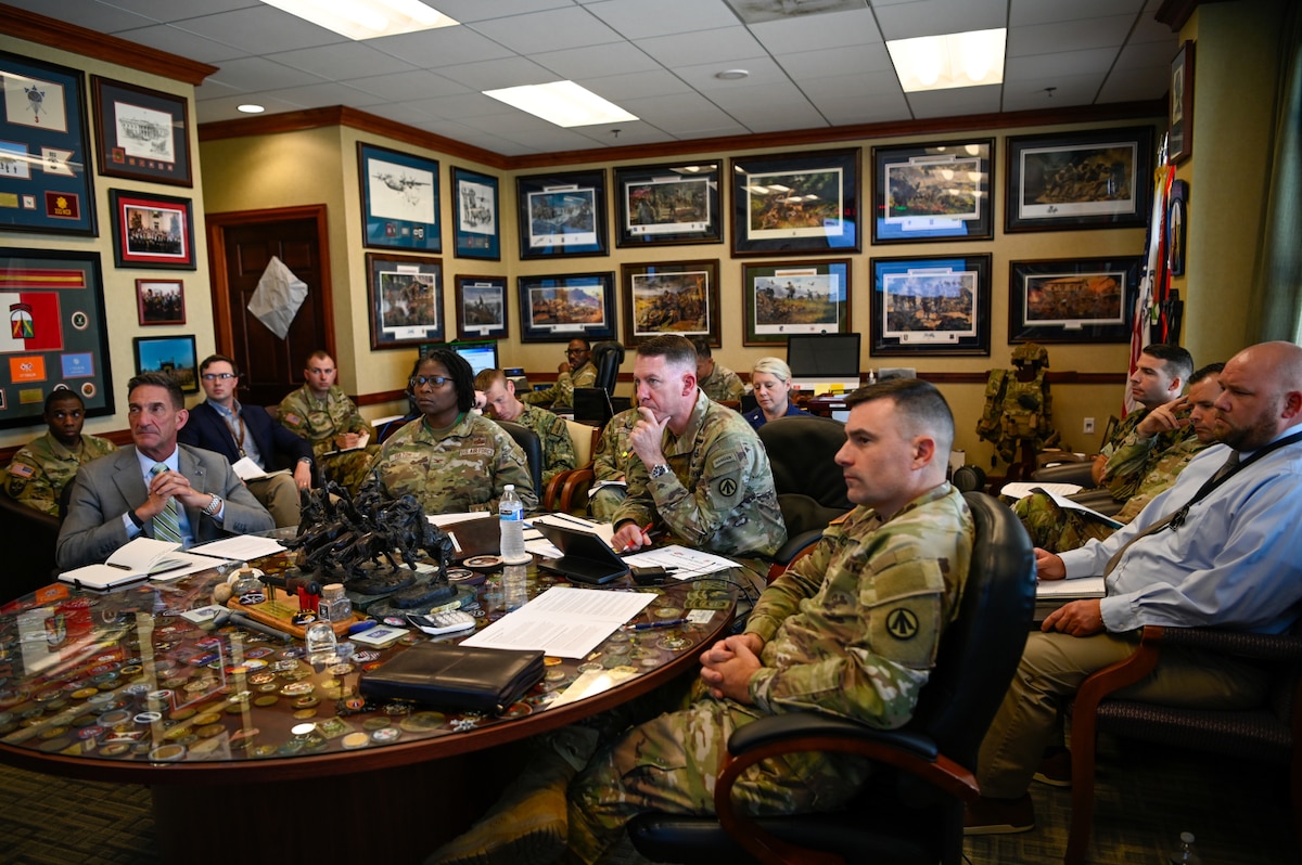 A group of people wearing camouflage military uniforms and business attire sit around a table and listen to a person speaking who is not in the frame, while others in similar attire sit in chair, along the walls. The walls are adorned with awards and pictures of military scenes.