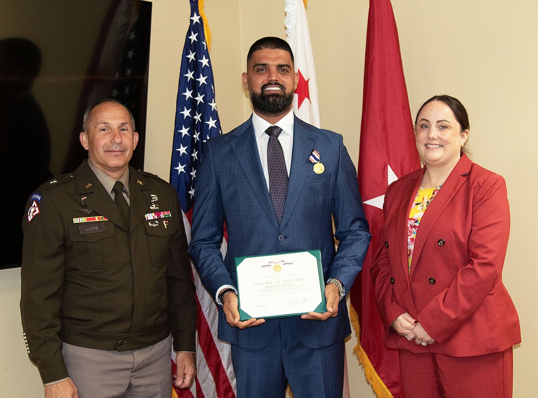 A man in a blue suite stands between a Soldier and a woman holding a certificate and has a medal pinned to his chest.