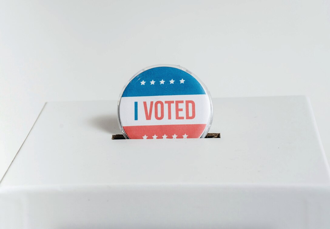 An "I Voted" button that is red, white and blue, is shown being dropped into a white ballot box.
