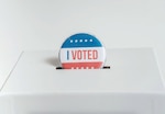 An "I Voted" button that is red, white and blue, is shown being dropped into a white ballot box.