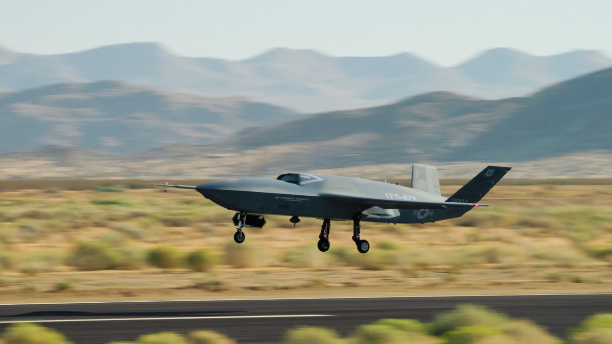 An unmanned military aircraft takes off from a runway and is currently about eight feet above the tarmac. There is a desert and mountains in the background.