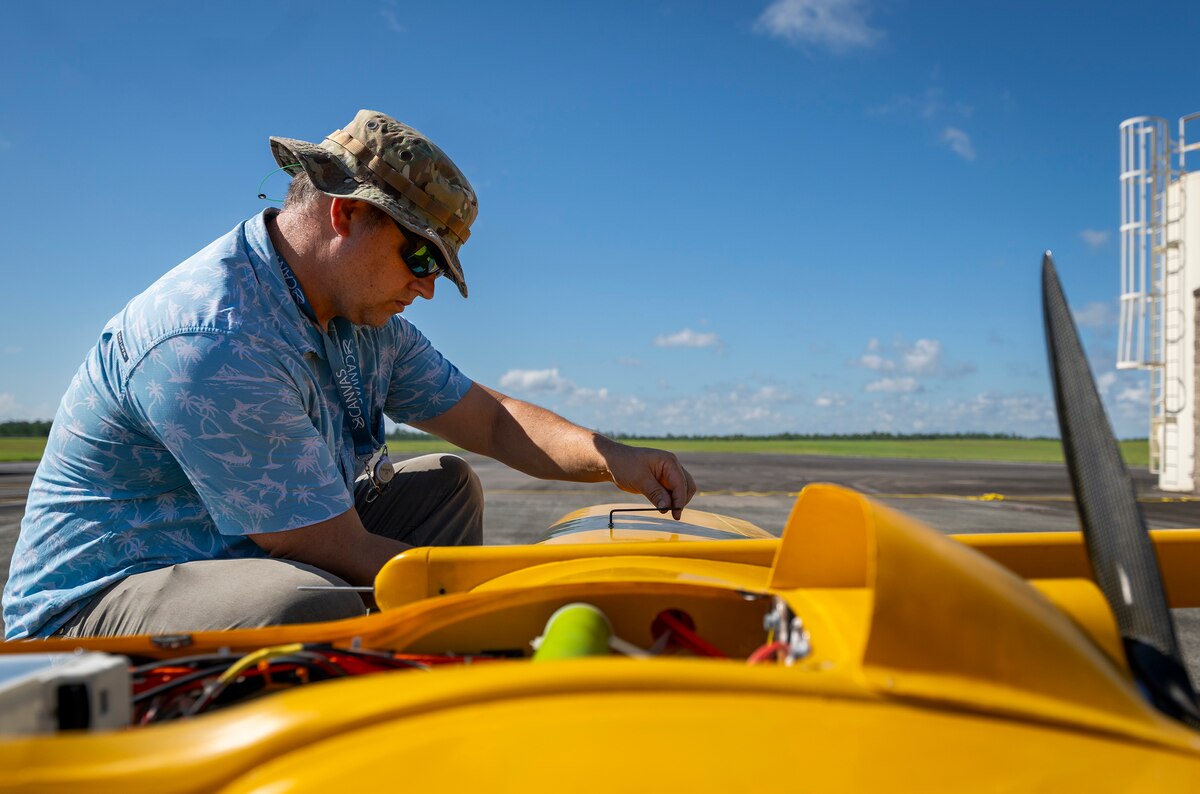 A man in casual attire and a camouflage hat tightens a hex wrench on a bright orange unmanned aircraft.