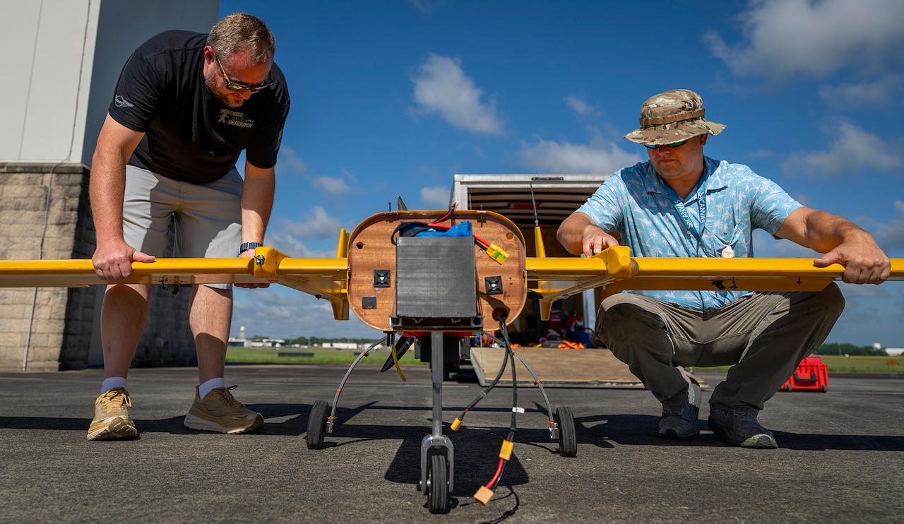 Two men in casual attire, one wearing a camouflage hat, work on a bright orange unmanned aircraft on a tarmac. The front of the aircraft is exposed with wires hanging down and there is an open trailer in the background.