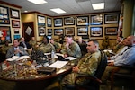 A group of people wearing camouflage military uniforms and business attire sit around a table and listen to a person speaking who is not in the frame, while others in similar attire sit in chair, along the walls. The walls are adorned with awards and pictures of military scenes.