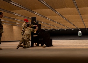 A military member crouches down with a handgun behind a wooden barrier with square holes cut out.