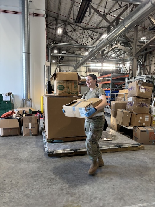 A woman wearing gloves, camouflage pants and a T-shirt, carries a box. There are several boxes stacked in the background.