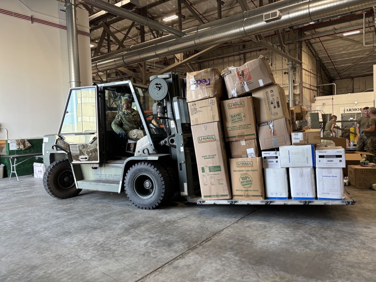 A man wearing a camouflage military uniform drives a forklift loaded with boxes. There are three other people in similar attire standing in the background.