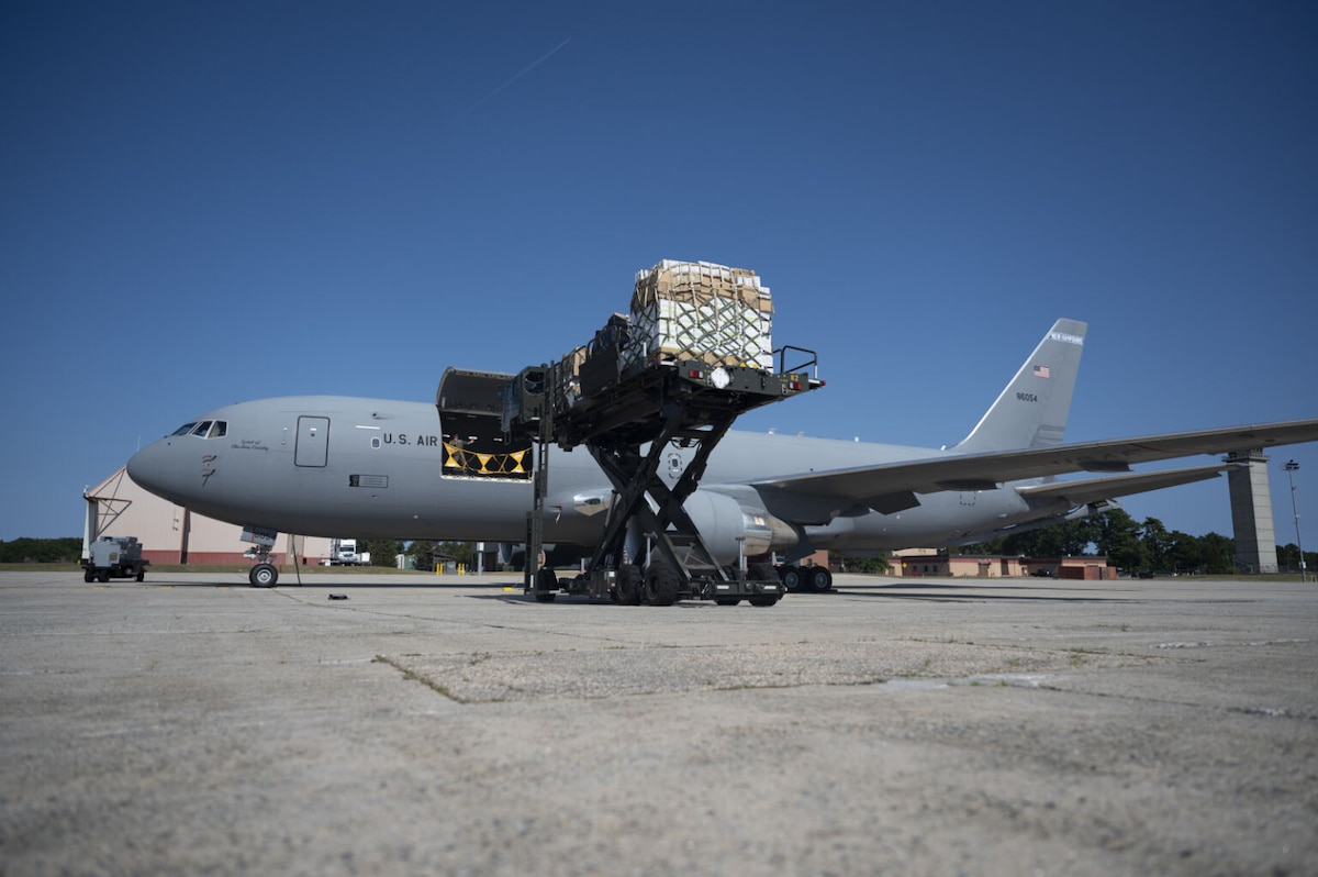 A large gray military cargo aircraft is parked on a tarmac with a cargo bin open. A pallet of supplies is on a loader to the side of the aircraft opening.