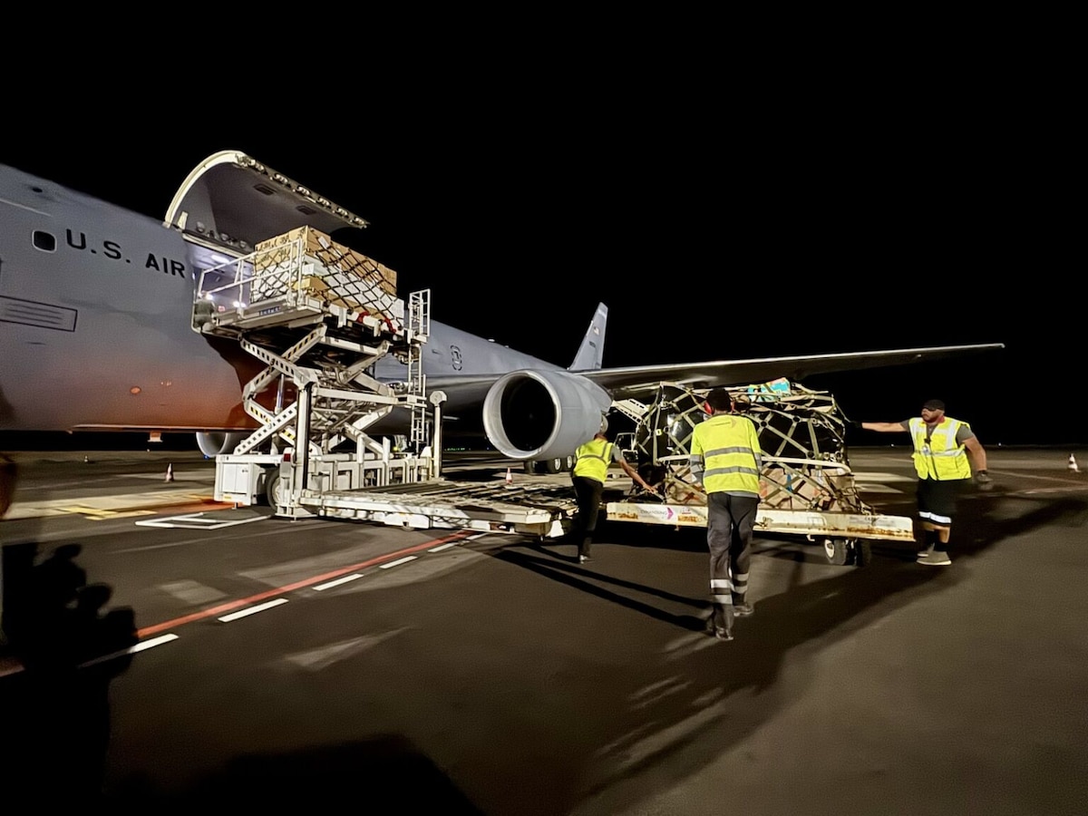 Three men wearing civilian attire and yellow reflective vests offload cargo from a large gray military aircraft.