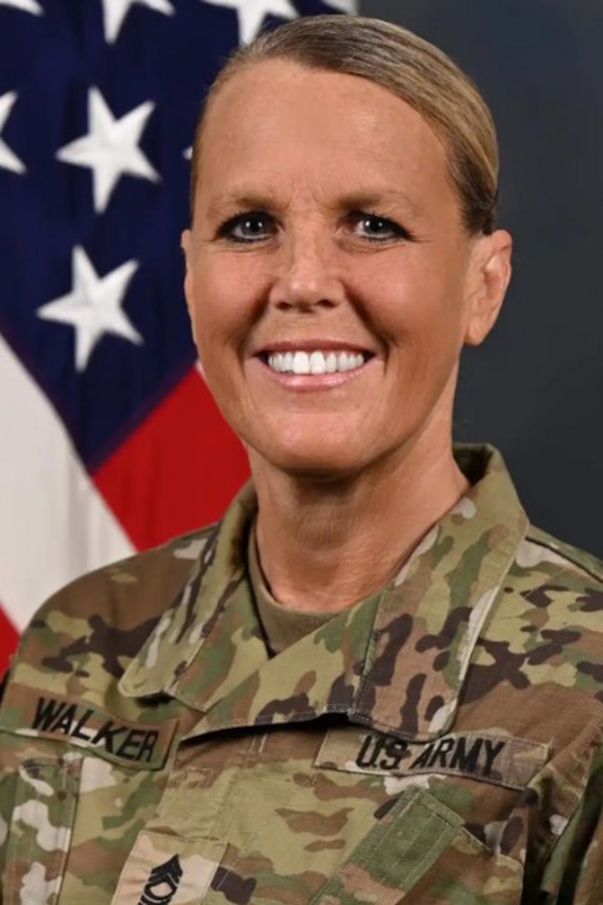 A person wearing a camouflage jacket smiles and poses in front of an American flag.