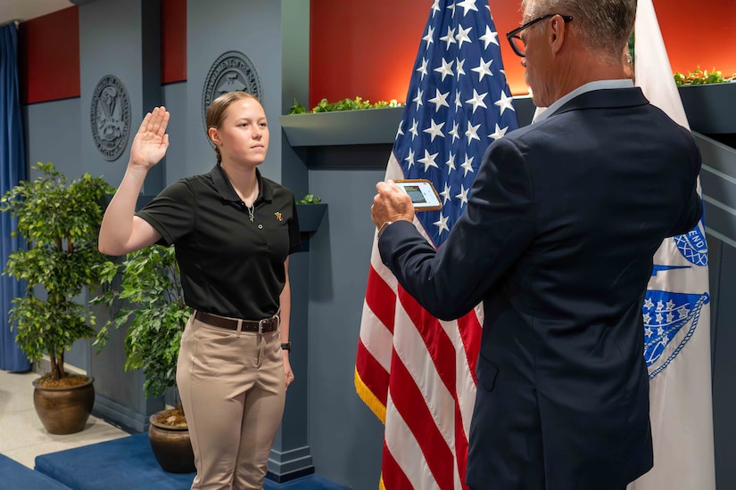 A person raises their right hand and faces another person in a suit while standing in front of flags instead a blue and red room.