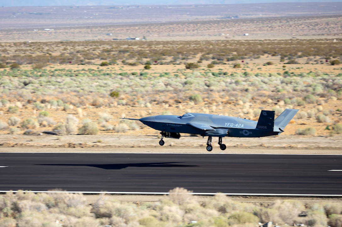 A small grey aircraft flies just above a runway surrounded by arid terrain.