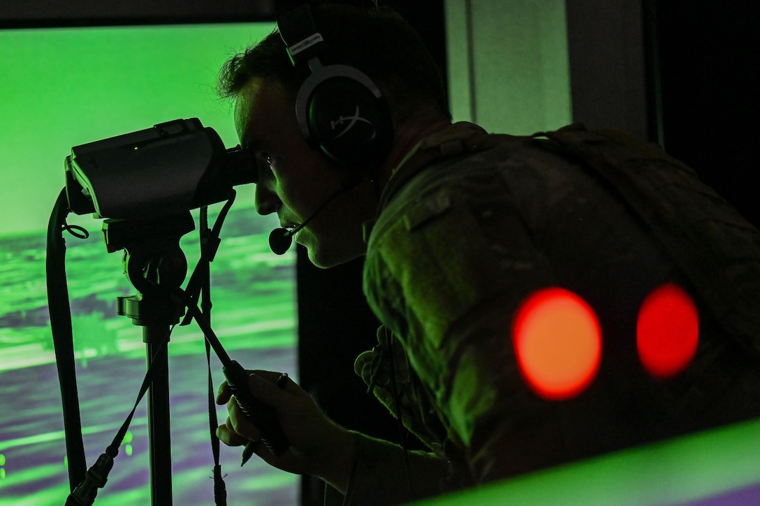 An airman wearing a headset looks through a simulator in front of a screen illuminated by a greenish color with two red circles.