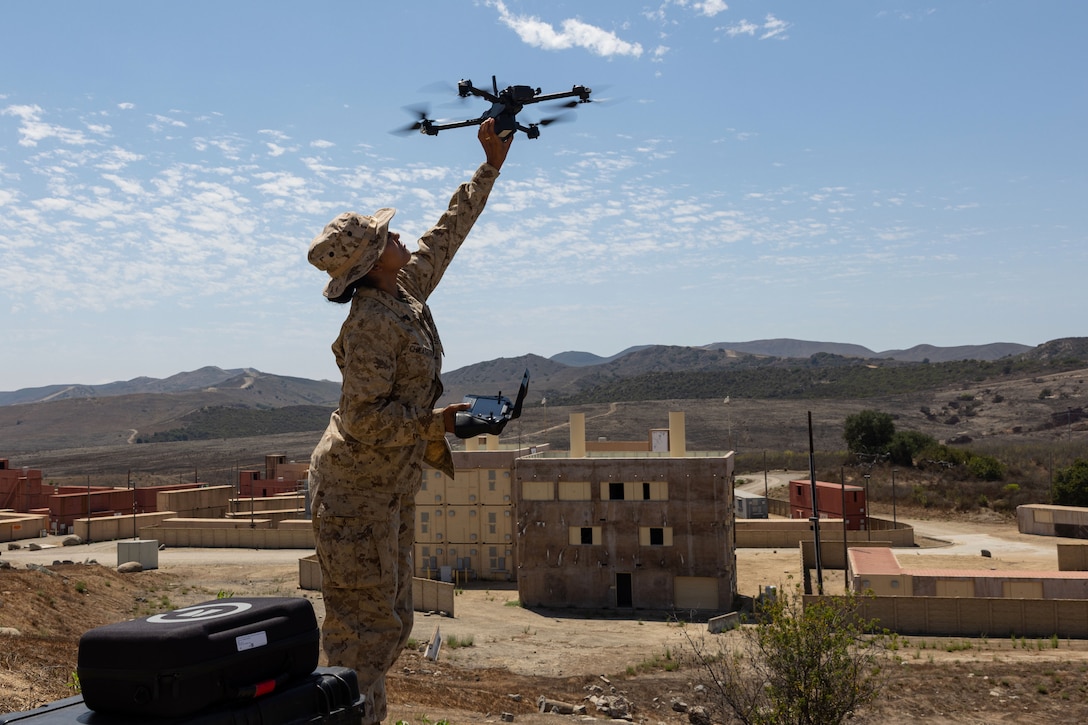 A Marine retrieves a drone from a blue sky with concrete structures and mountains in the background.