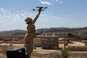 A Marine retrieves a drone from a blue sky with concrete structures and mountains in the background. 