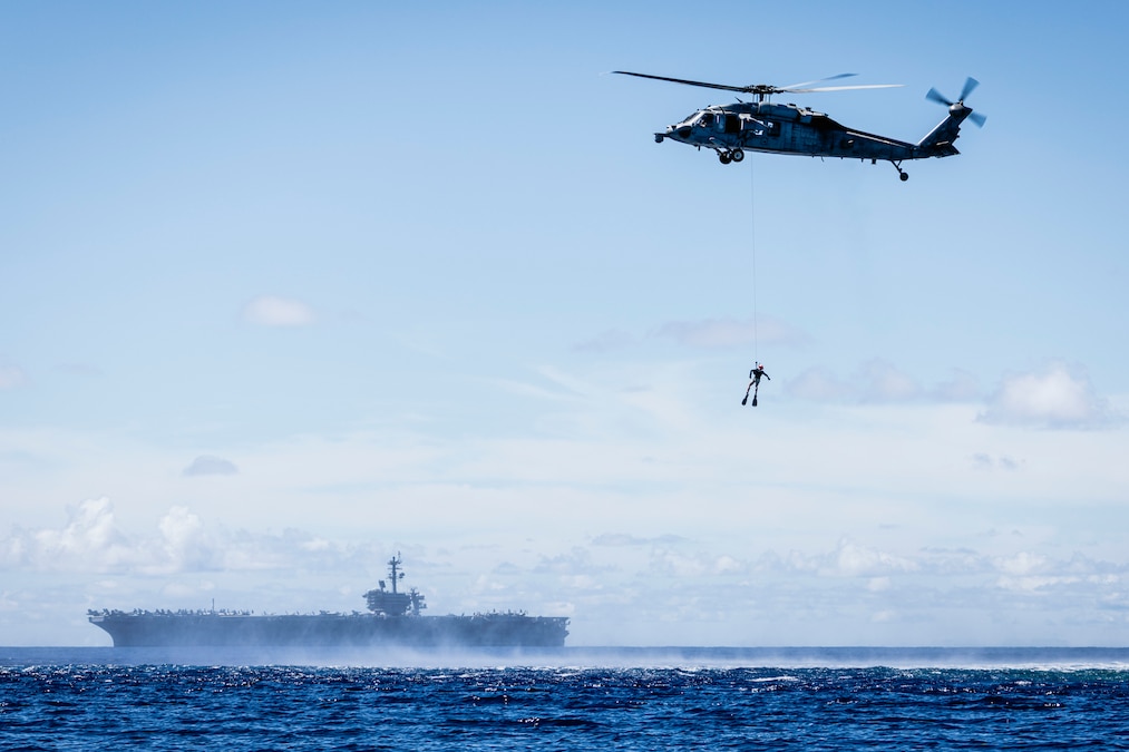 A sailor is hoisted to a hovering helicopter as a ship transits a body of water during the day.