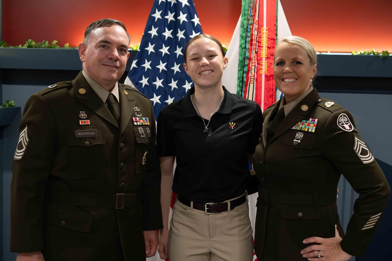 Three service members stand together and pose for a picture in front of two standing flags inside a room.