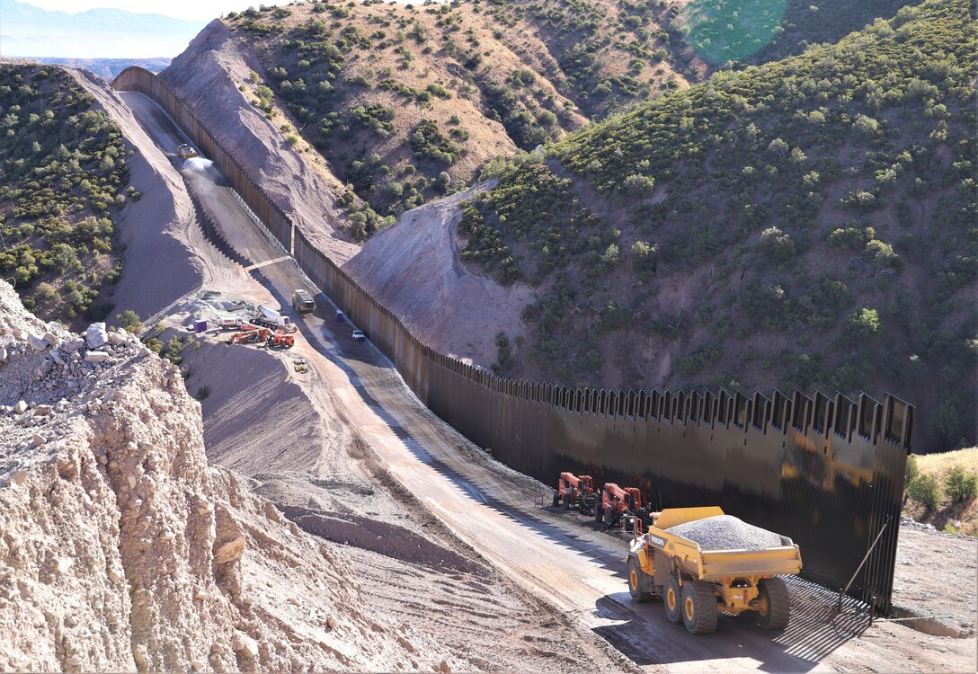 Construction vehicles travel down a dirt road next to newly constructed sections of barrier fencing.