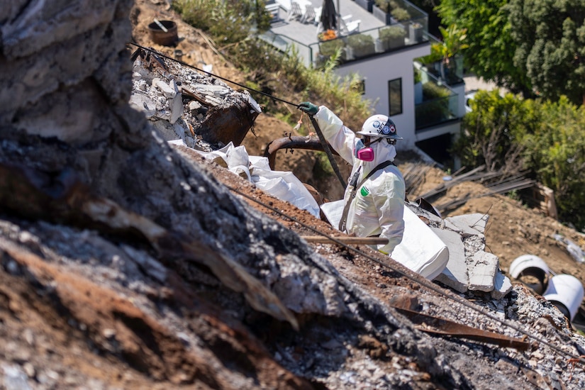 A person wearing a protective suit, helmet and respirator, uses a rope to move along a rugged slope to collect debris left by a wildfire.