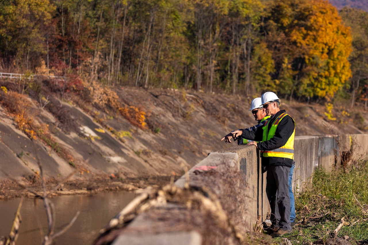 Two men wearing civilian attire, helmets and reflective vests stand at an embankment of a flood channel. There are trees across the channel in the background.