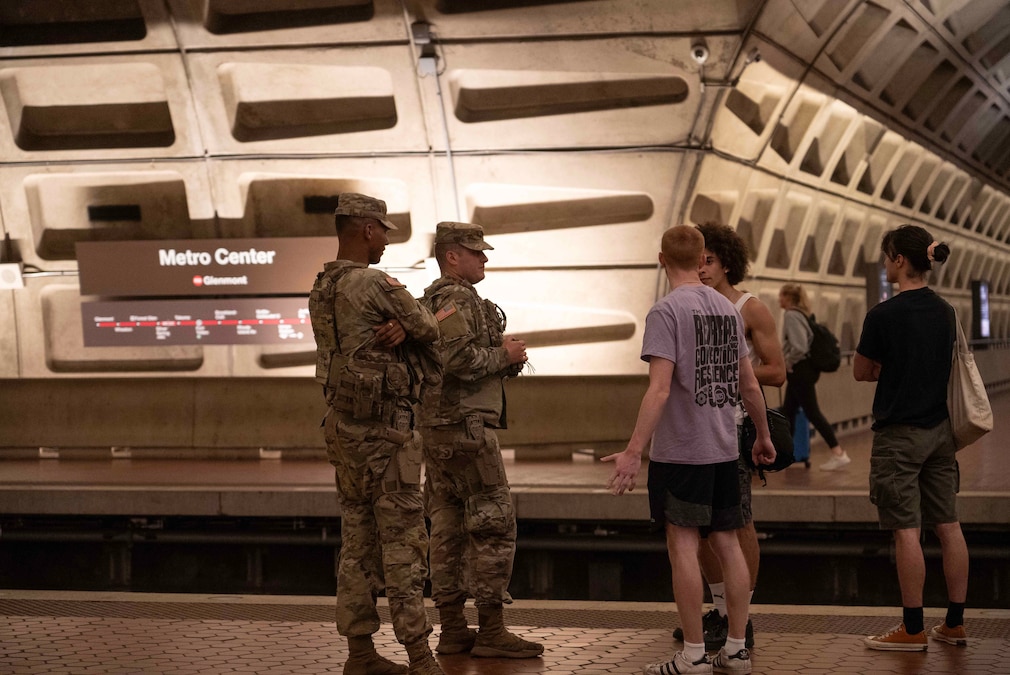 Two guardsmen speak to three people while standing next to train tracks inside a dimly lit station with a sign behind that says, "Metro Center."