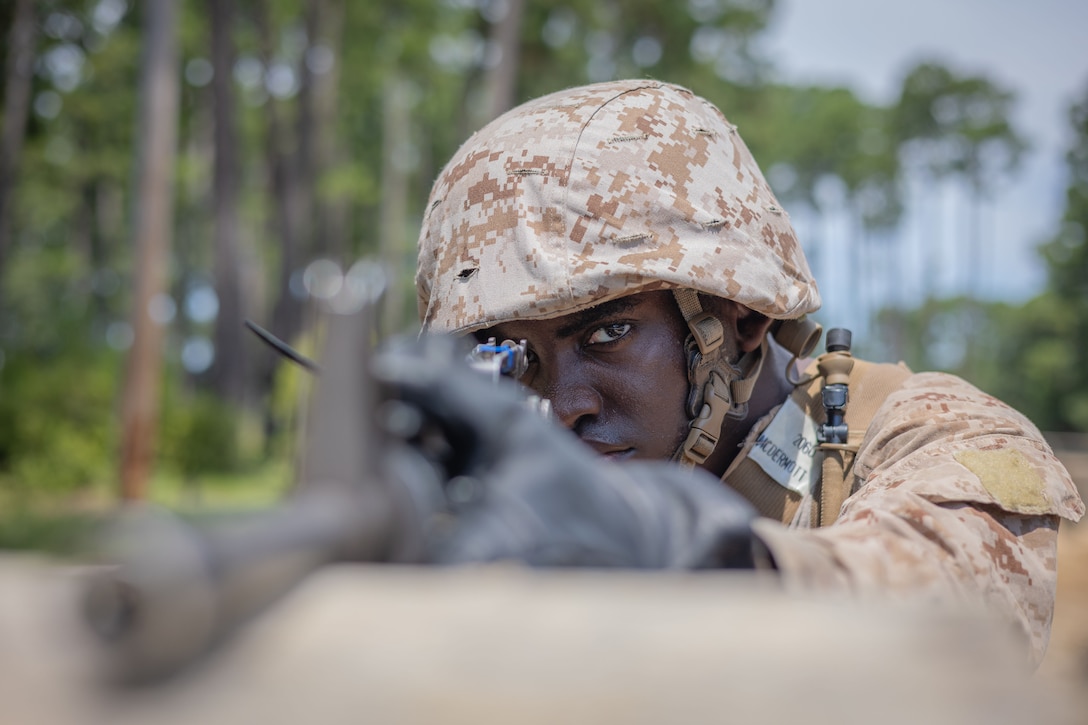 A Marine Corps recruit in a helmet aims a service weapon outside in front of a forest, as seen from head-on.