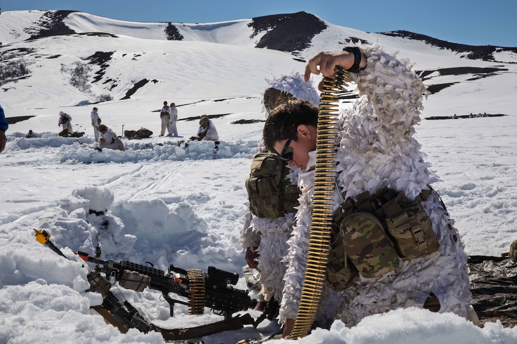 Two soldiers in snow camouflage load an automatic weapon while kneeling in snow during the day while other soldiers stand in the distance in front of snowy hills.