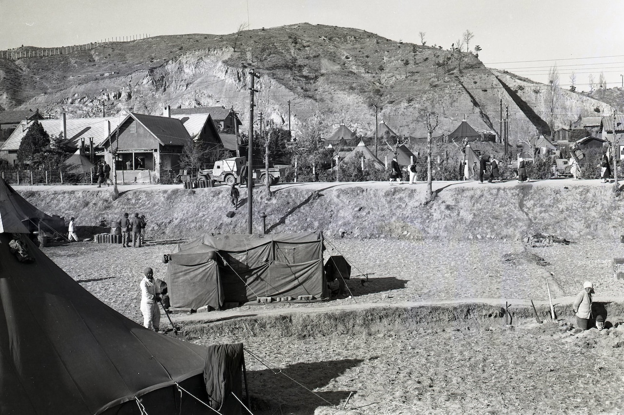 A small building and some tents are set up beside a barren hill. There are people in civilian attire walking around the area, and large buildings in the background.