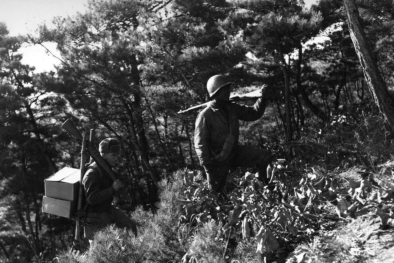 Two men in military uniforms carrying guns and boxes march up a hillside.