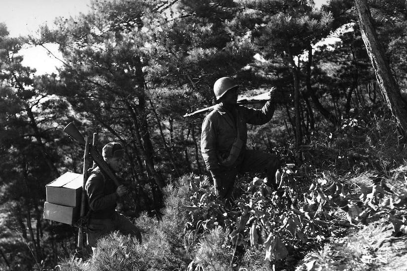 Two men in military uniforms carrying guns and boxes march up a hillside.