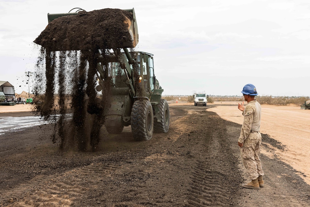A construction vehicle picks up dirt on a dirt road while a Marine in a helmet directs from the right.