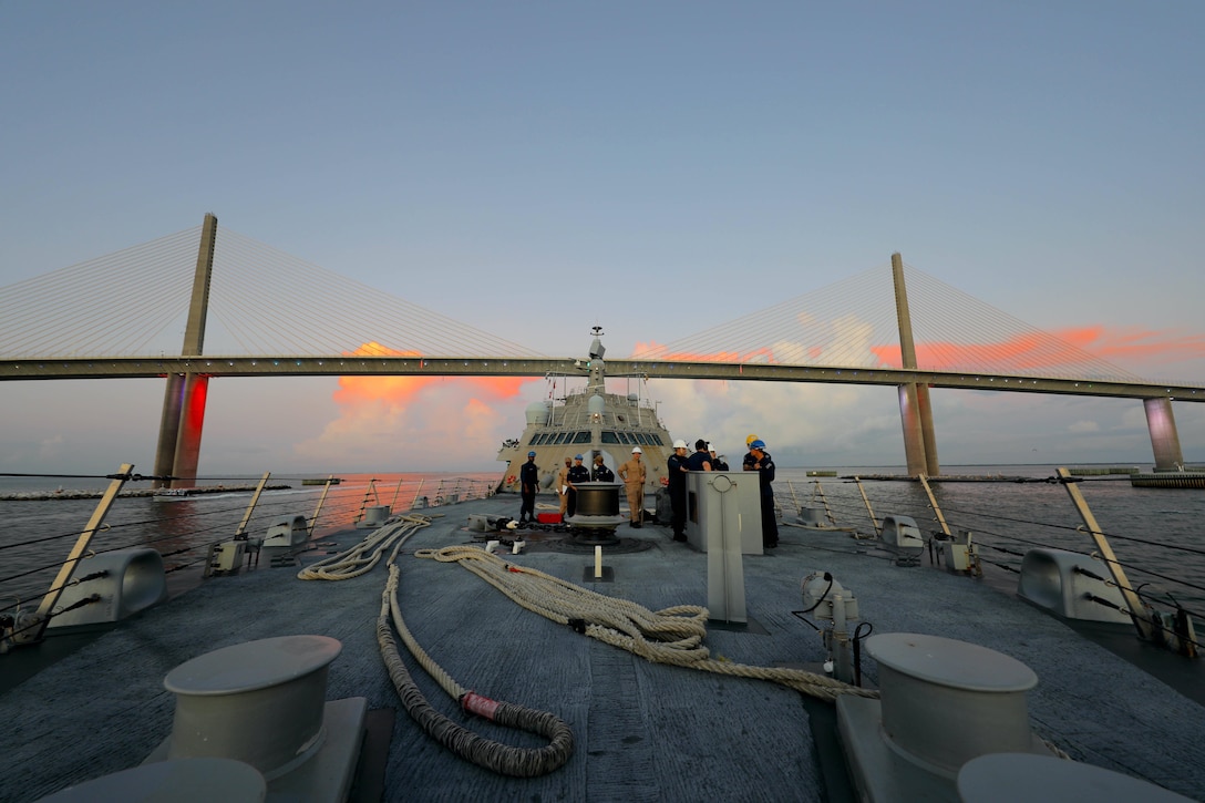 A group of sailors stand on the deck of a ship as it passed under a bridge with orange clouds under a blue sky.