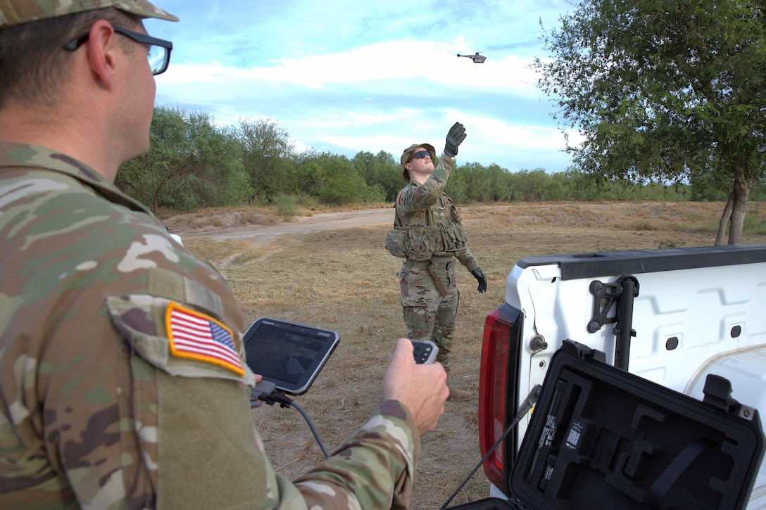 A soldier holds drone equipment next to a truck and watches another soldier reach up towards a drone in flight outside.
