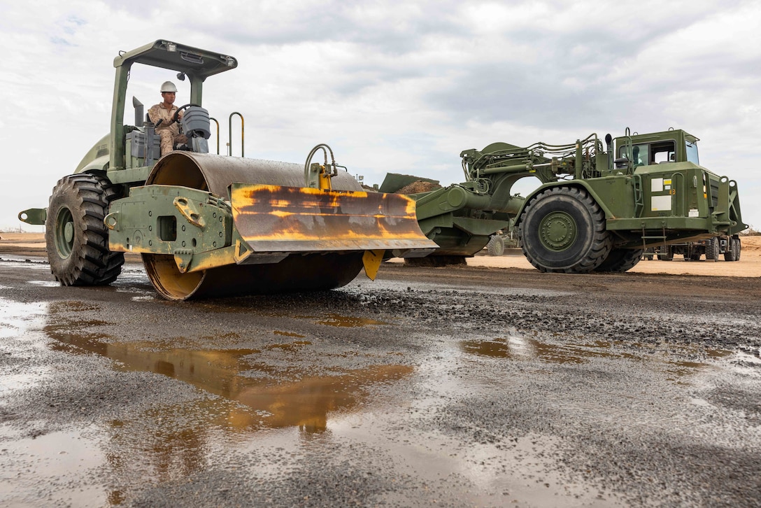 A soldier drives a green tractor on a slick road while another green tractor is parked behind to the right on a cloudy day.