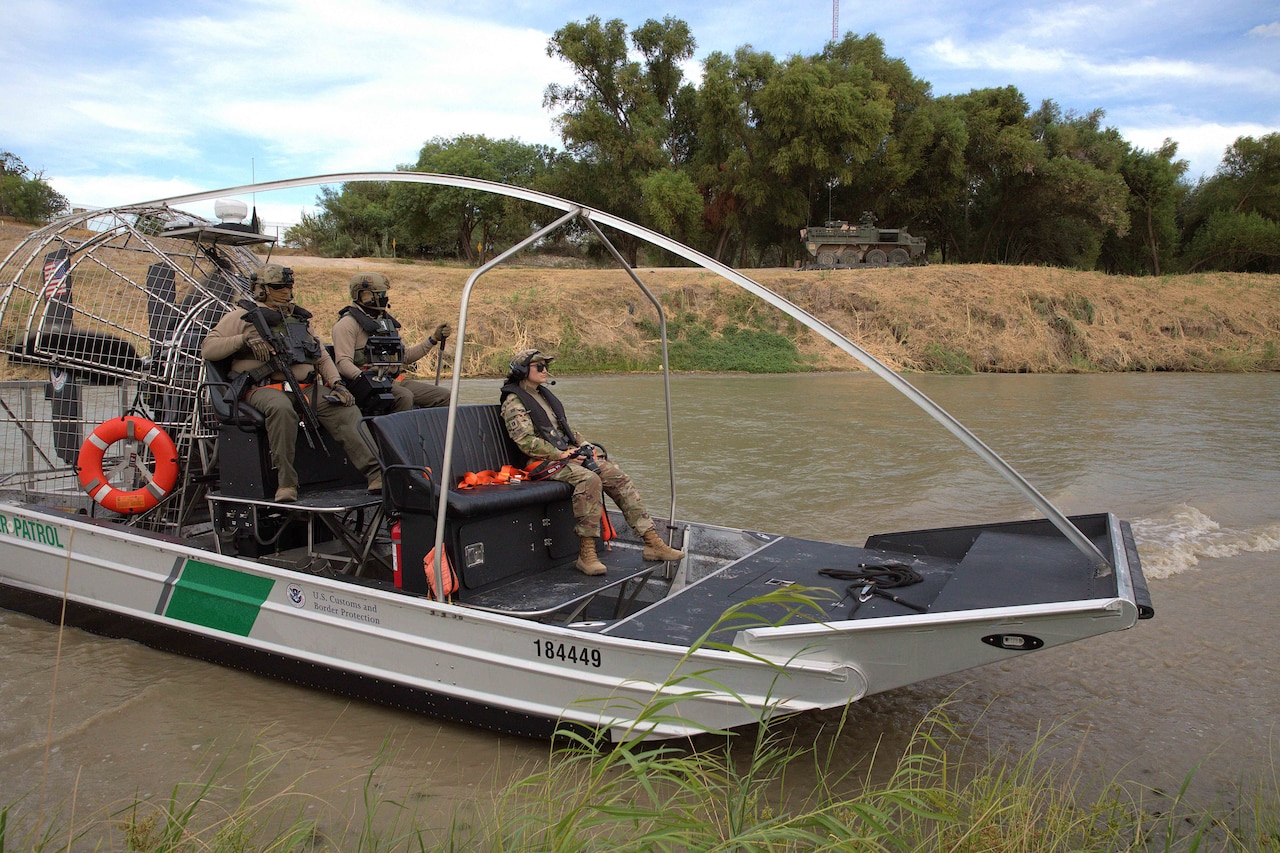 Three people sit in a metal boat on a small body of water under blue skies with trees and tall grass in the background.