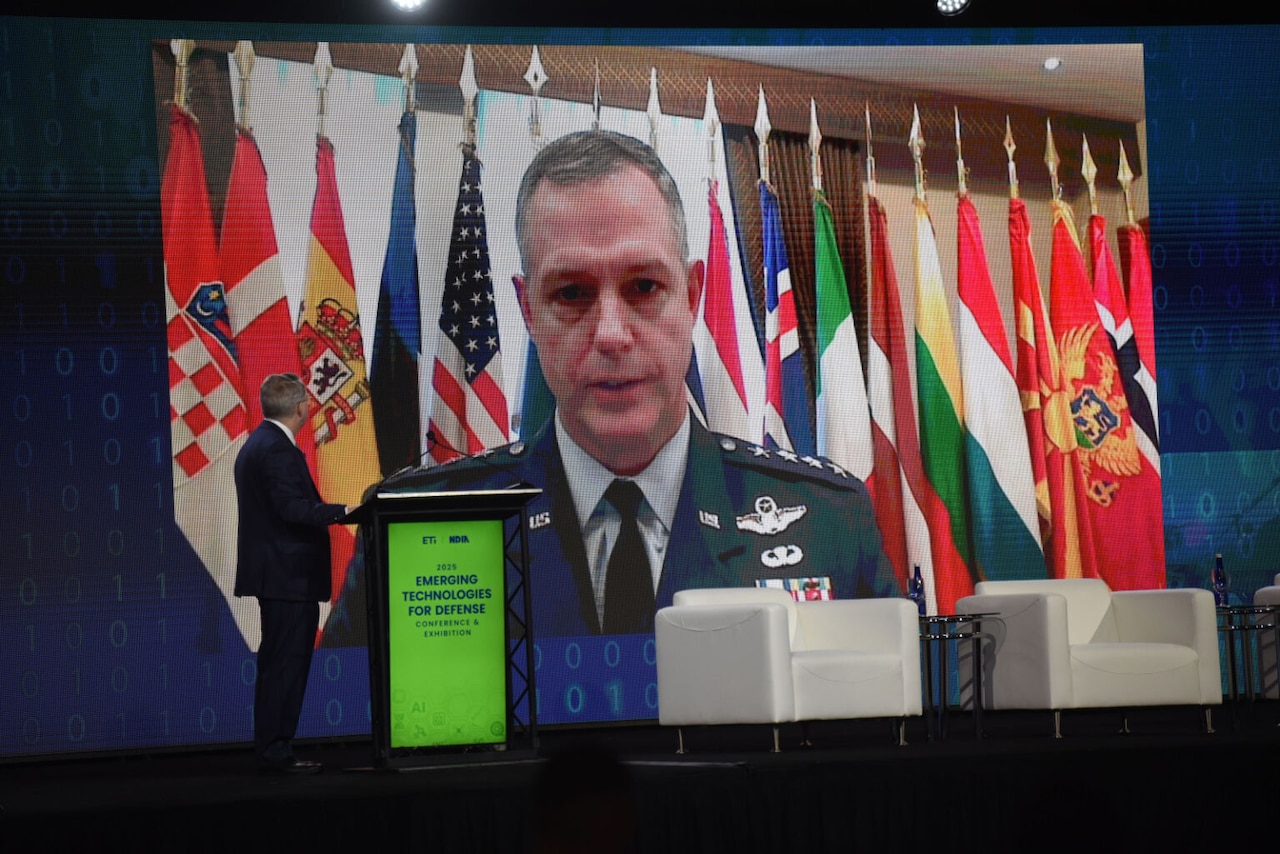 A man in a military dress uniform speaks on a big screen with flags in the background. There is a man in business attire standing by a podium with his back to the camera in the foreground.