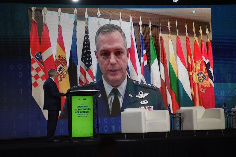 A man in a military dress uniform speaks on a big screen with flags in the background. There is a man in business attire standing by a podium with his back to the camera in the foreground.