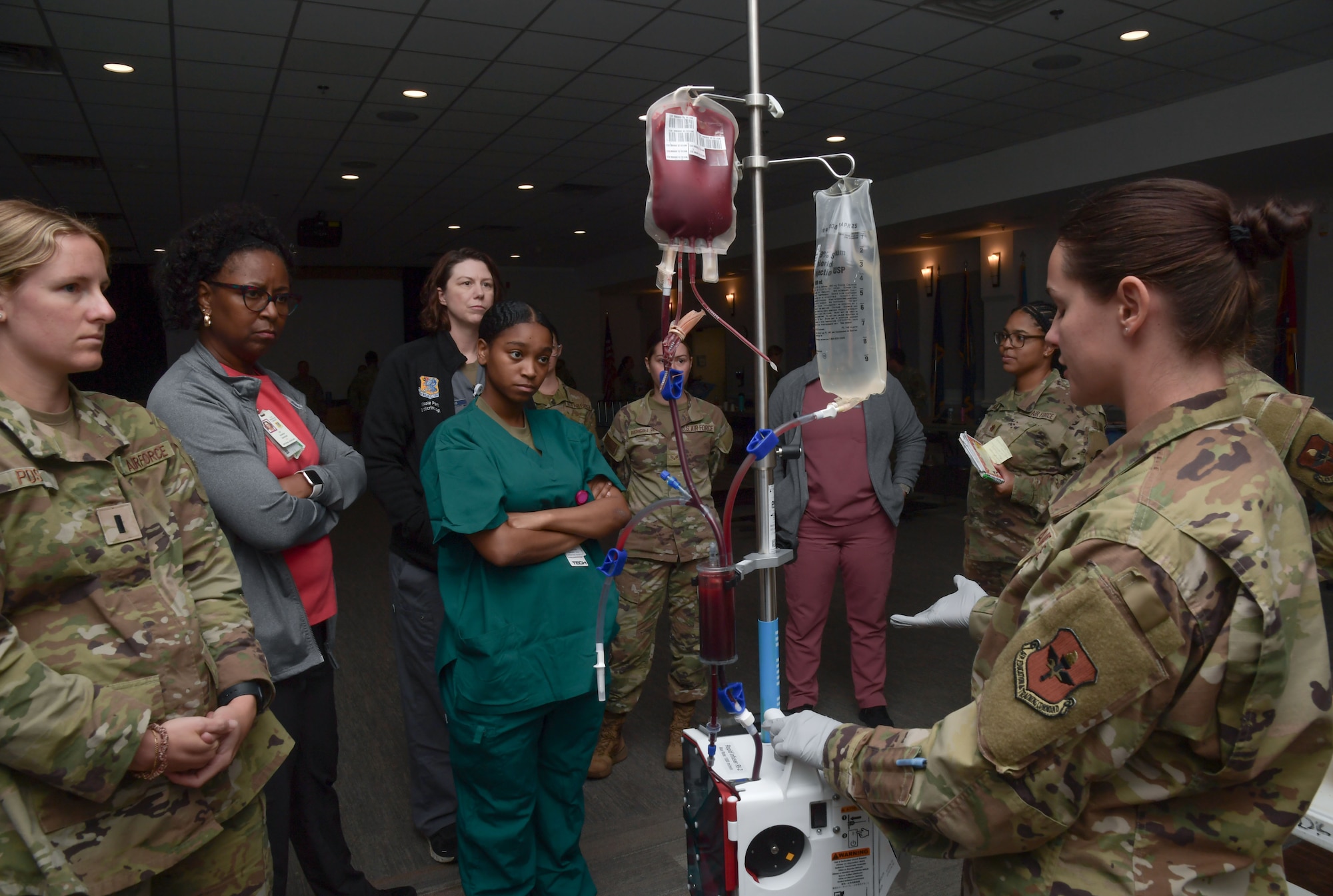 A woman in a military uniform speaks to a group of people standing next to an IV stand.
